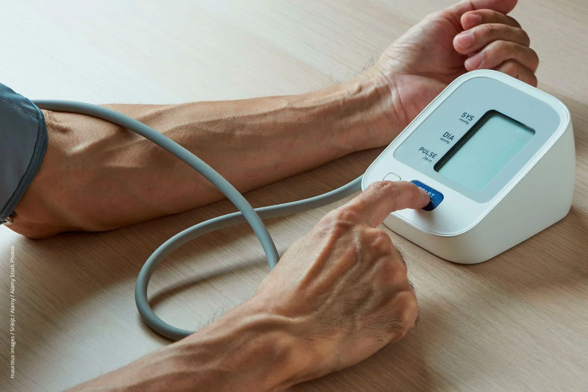 
Mann sitzt an einem Tisch und misst sich mit einem Blutdruckmessgerät selbst den Blutdruck.