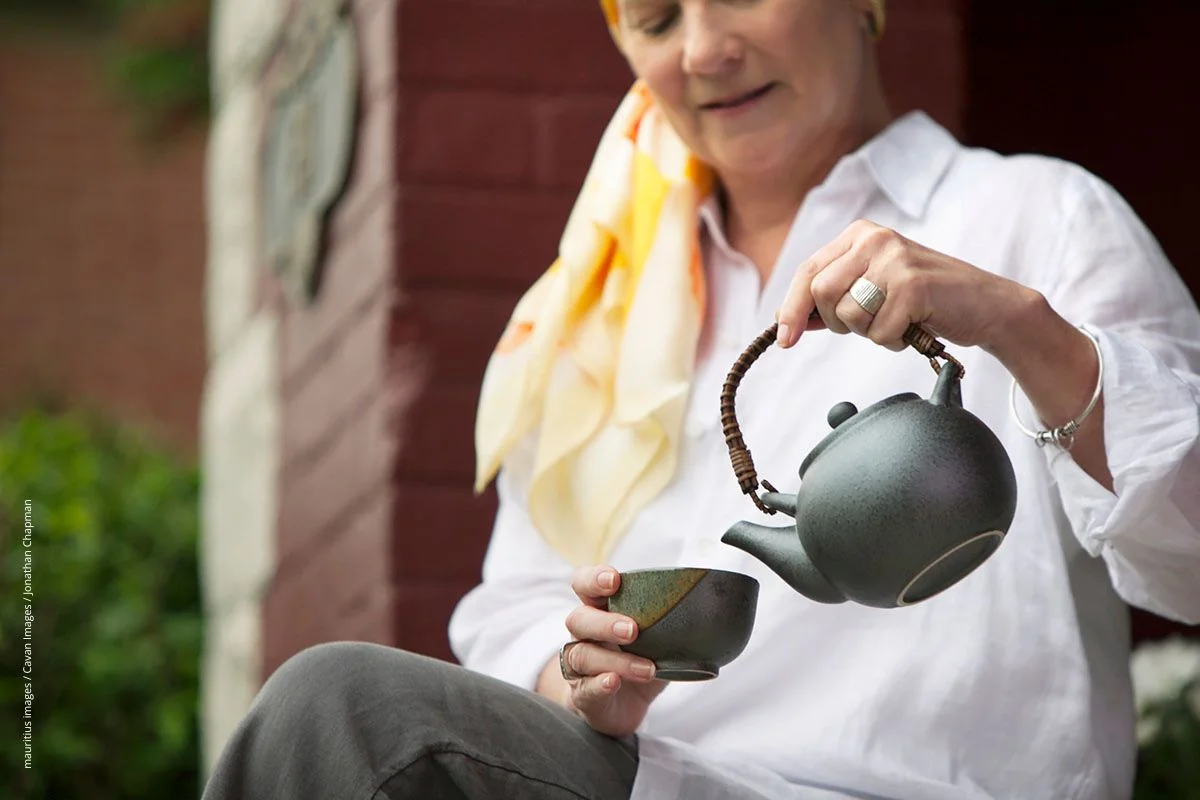 Frau in mittleren Jahren gießt sich Tee aus einer Kanne ein. Teetrinken hat einen guten Ruf für die Gesundheit.