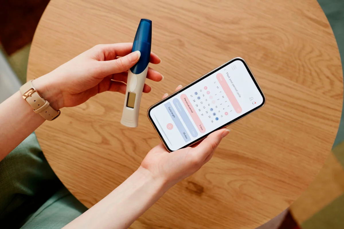 Eine Frau hält einen Schwangerschaftstest Hand, in der anderen einen Kalender auf dem Smartphone.