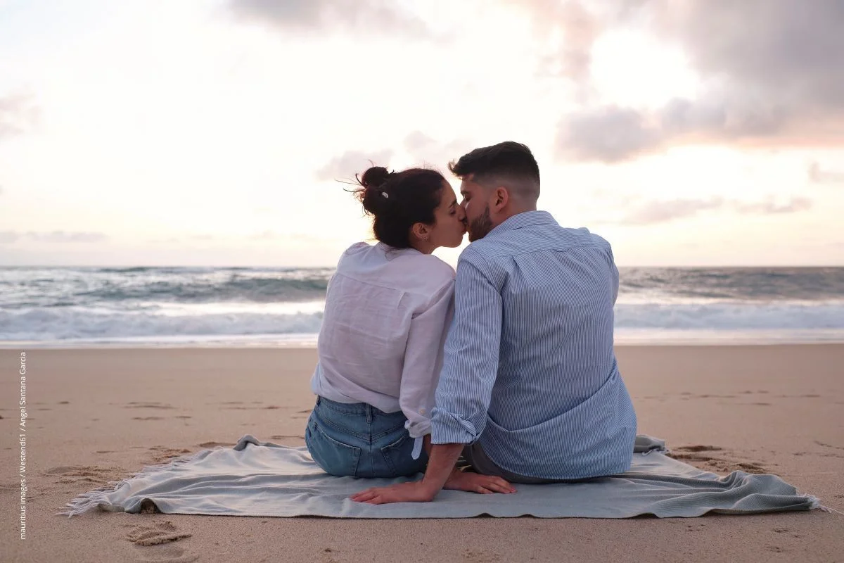 Ein Pärchen sitzt bei Sonnenuntergang am Strand und küsst sich.