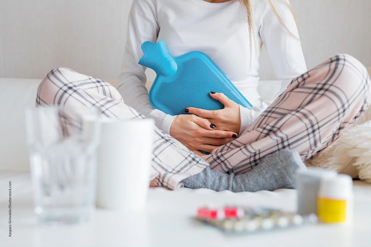 Sitzende Frau hält eine Wärmflasche an ihren Bauch. Im Vordergrund sind Wasserglas, Teebecher und Tabletten zu sehen. Wärme kann gegen Bauschchmerzen helfen.
