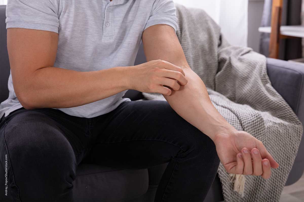 Mann sitzt auf dem Sofa und kratzt sich am Arm. Großflächiger und anhaltender Juckreiz sollte ärztlich abgeklärt werden.